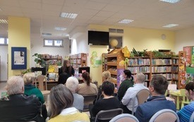 Starszy mężczyzna przemawia do zgromadzonej publiczności, w tle regały z książkami.