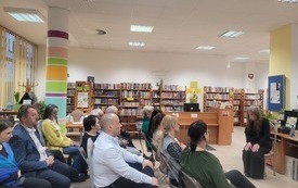 Grupa ludzi siedzi na krzesłach, w tle regały z książkami