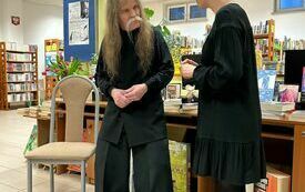 Mężczyzna z wąsami i długimi włosami stoi obok młodej kobiety ubranej na czarno, w tle regały z książkami.