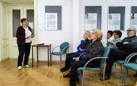 Starsza kobieta przemawia do zebranych w sali osób.