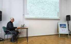 Przy stoliku z laptopem siedzi starszy mężczyzna, na ścianie prezentacja multimedialna