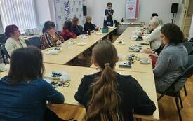 Uczestnicy warsztatów siedzą przy stołach, przed nimi prowadząca spotkanie kobieta.