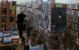 Dzieci wchodzą za bibliotekarką pomiędzy regały z książkami