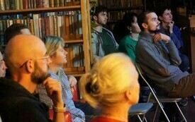 Grupa osób siedzących pomiędzy bibliotecznymi regałami i patrzących w tym samym kierunku