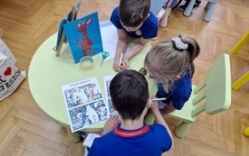 Okragły stoliczek i tr&oacute;jka dzieci rozwiązujących zadania. 