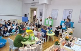 Przy dw&oacute;ch małych stolikach siedzą dzieci, bibliotekarka prowadzi turniej. W tle drużyny kibicujące. 
