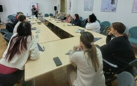 Grupa kobiet siedzi przy stole i patrzy na kobiete prowadzącą warsztaty