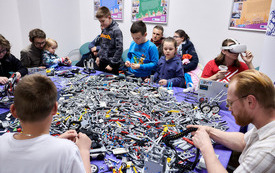 Stół z klockami LEGO otoczony dziećmi i dorosłymi budującymi.