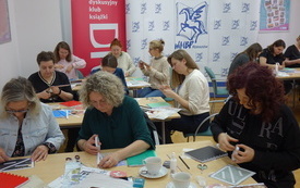 Bibliotekarki przy stolikach tworzą prace z materiał&oacute;w papierniczych. 