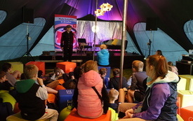 Pod namiotem grupa dzieci ogląda występ młodego mężczyzny w kapeluszu