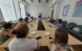 Przy bardzo dużym stole siedzą kobiety i słuchają prowadzącej.