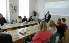 Mężczyzna w marynarce przemawia do zgromadzonych na naradzie osób.