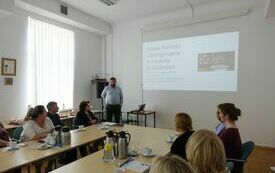 Mężczyzna w niebieskiej koszuli przemawia do uczestników narady.