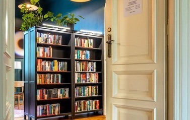 Wnętrze biblioteki sfotografowane przez uchylone drzwi. W centrum kadru znajdują się dwa czarne regały wypełnione książkami.